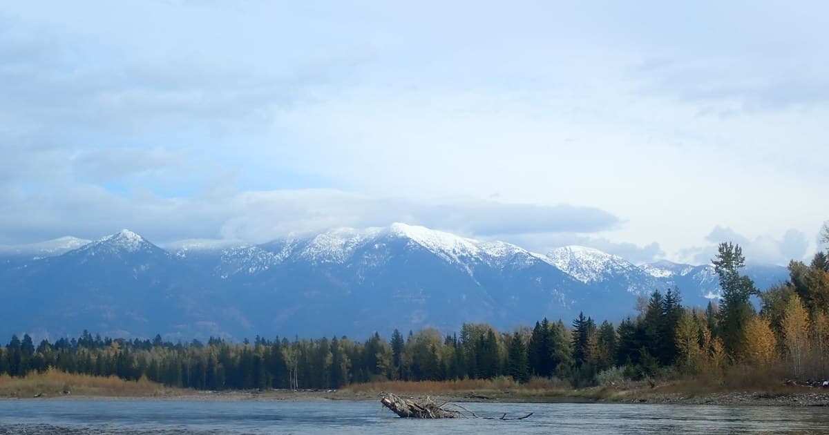 Peaceful view of Whitefish River with snow-capped mountains and autumn forest in Whitefish, Montana.